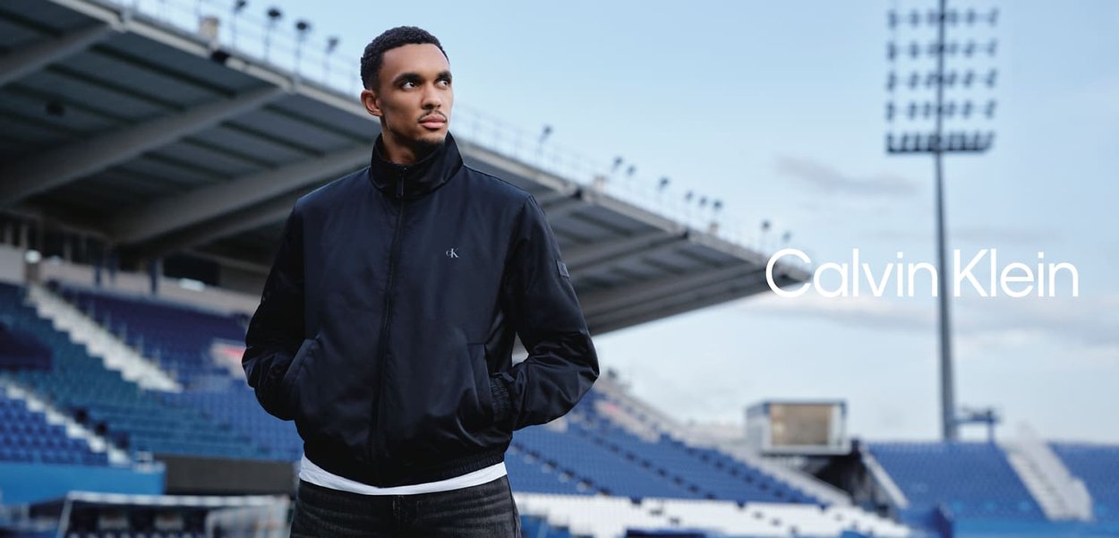 Mann i sort Calvin Klein-jakke med hendene i lommene som står i en tom idrettsstadion under flombelysning.