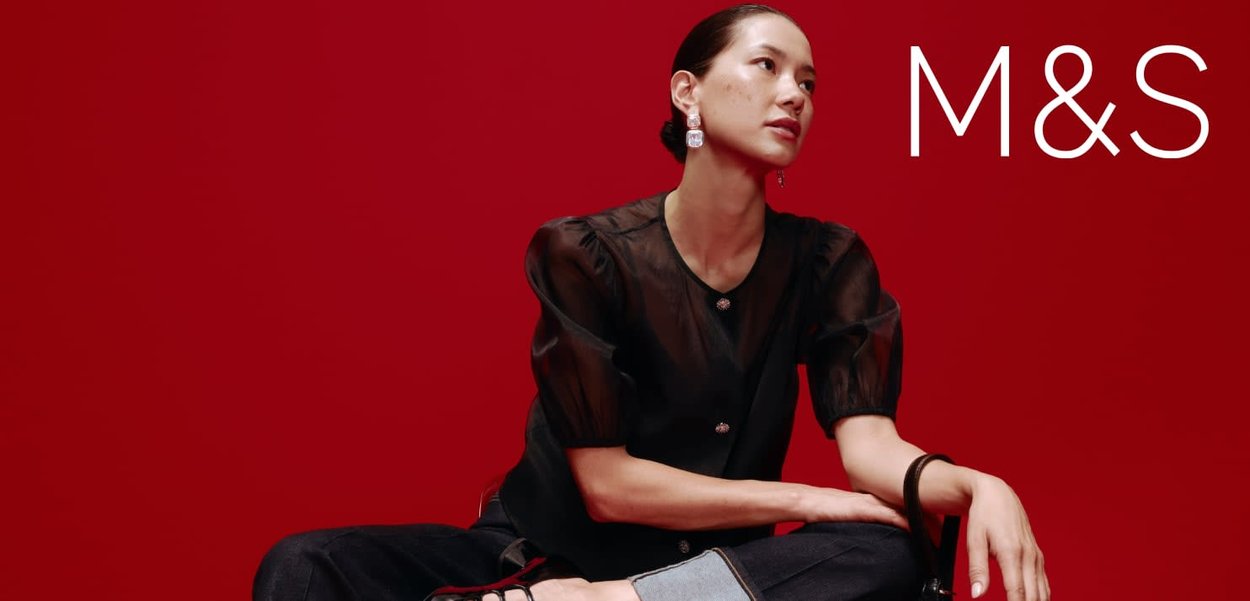 Blouse noire transparente à manches bouffantes, détails à boutons ; jean foncé taille haute ; modèle assis devant un fond rouge. Logo « M&S » en blanc.