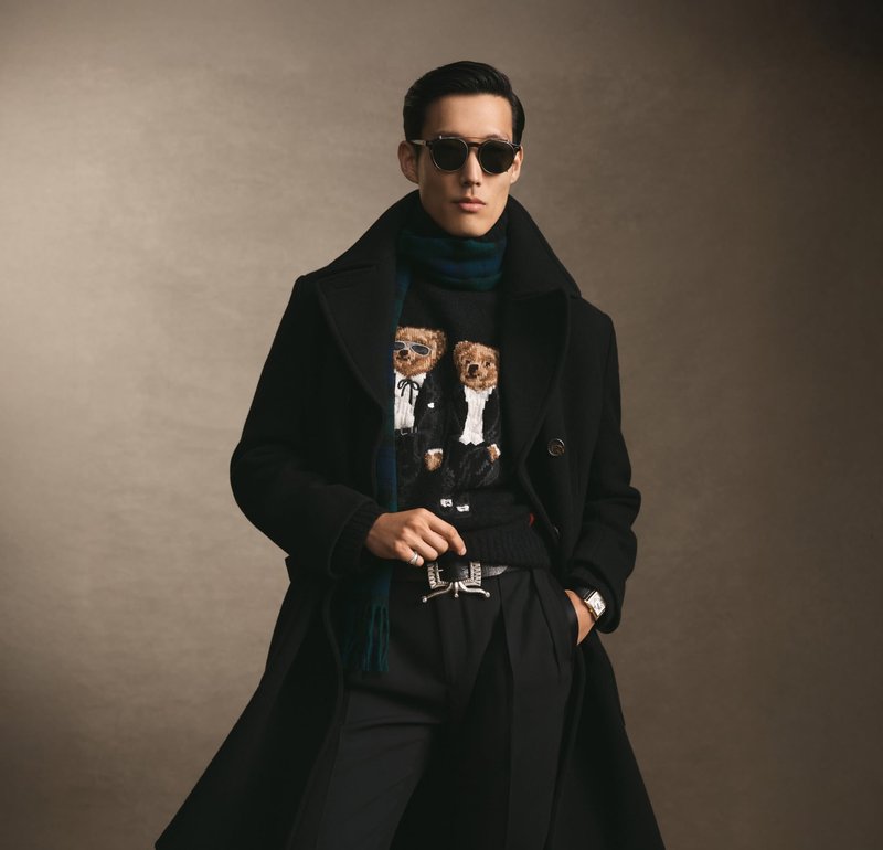 Manteau en laine noire avec larges revers, pull à motif présentant deux ours en smoking, pantalon noir et lunettes rondes. Montre argentée visible.