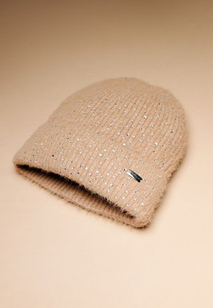 Beige Strickmütze mit einer weichen, flauschigen Textur, verziert mit silbernen dekorativen Nieten und einem kleinen schwarzen Etikett am Rand.