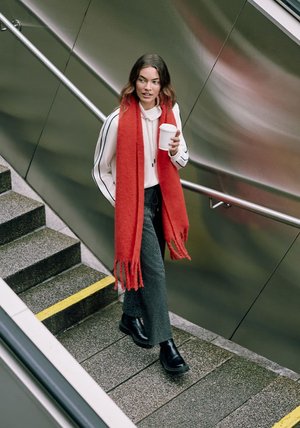 Czerwona szal, biała bluza z kapturem, wzorzyste spodnie i czarne botki za kostkę. Schodząc po betonowych schodach, trzyma filiżankę kawy w dłoni i metalową poręcz.