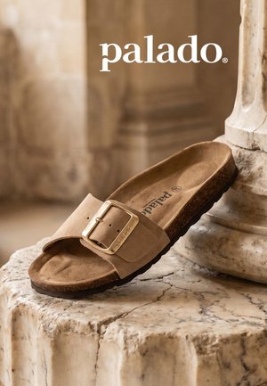 Beige suede slide sandal with large gold buckle and cork sole resting on textured marble surface with blurred stone background.