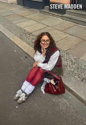 Une personne assise portant un gilet vert et grenat, un chemisier blanc, des collants rouges et des baskets argentées, à côté d'un sac à main rouge sur un sol pavé.