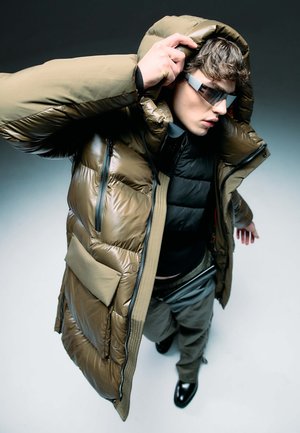 Gesteppten olivgrünen Parka mit hohem Kragen, Reißverschlüssen, Fronttaschen und glänzender Oberfläche. Kombiniert mit einer schwarzen Pufferweste und grauen Hosen.