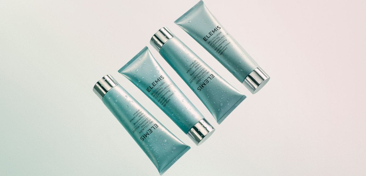 Fire blå Elemis Pro-Collagen rensende mussetuber med sølvkorker, arrangert diagonalt på en lys bakgrunn med vanndråper.