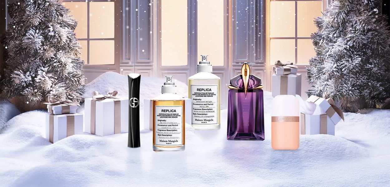 Collection of five perfume bottles on a snowy surface, with a frosted tree and gift boxes in the background, showcasing varied shapes and colors.