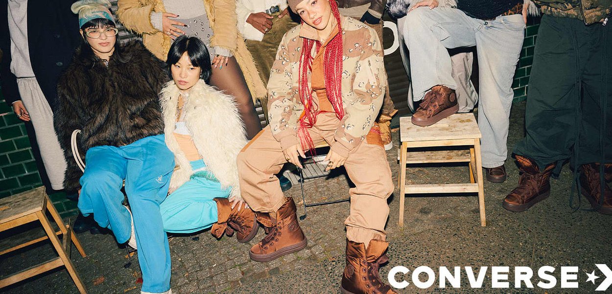 Tre persone con stili diversi: giacche oversize, pantaloni a fantasia e stivali marroni, sedute su sgabelli davanti a uno sfondo testurizzato.