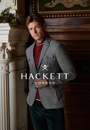 Grijze pied-de-poule blazer met zakdetails over een bordeauxrode trui, afgezet tegen een houten achtergrond, benadrukt door feestelijke groene accenten.