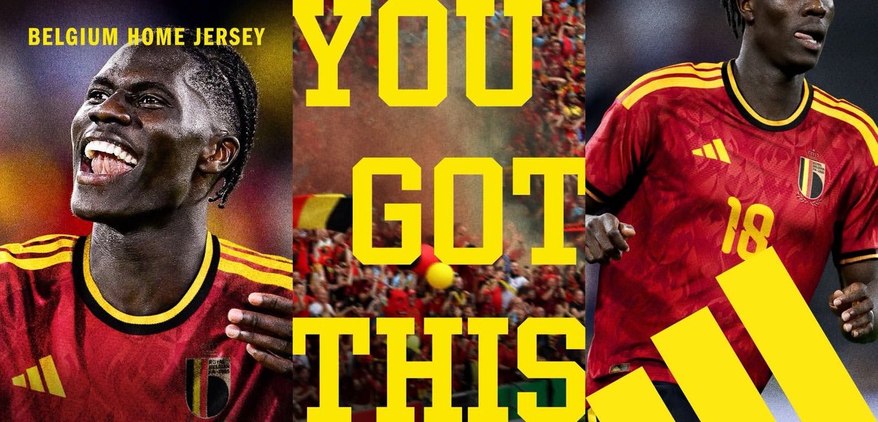 Maillot domicile rouge de la Belgique avec des accents jaunes et un logo couronne, présentant un motif texturé. Joueurs en train de célébrer, entourés de supporters.