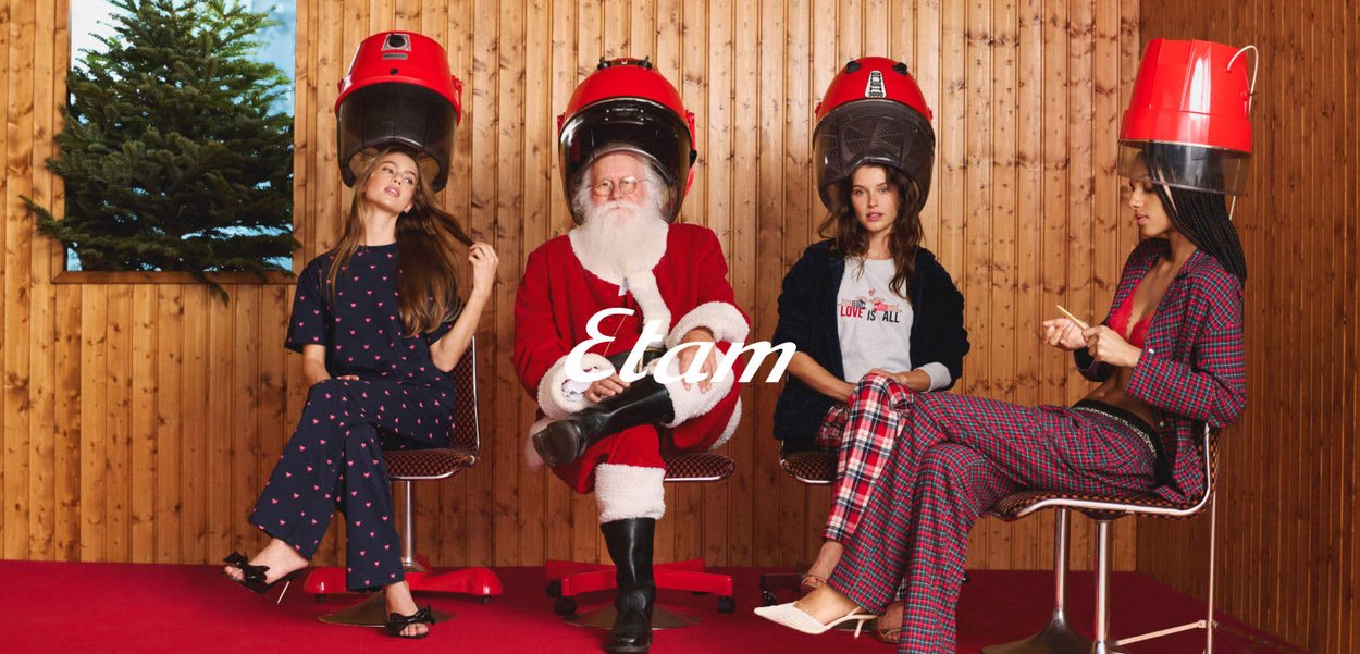 Quatre personnes dans un décor rétro, chacune sous un grand casque sèche-cheveux rouge. On y trouve des murs en bois, un tapis rouge et des tenues à motifs.