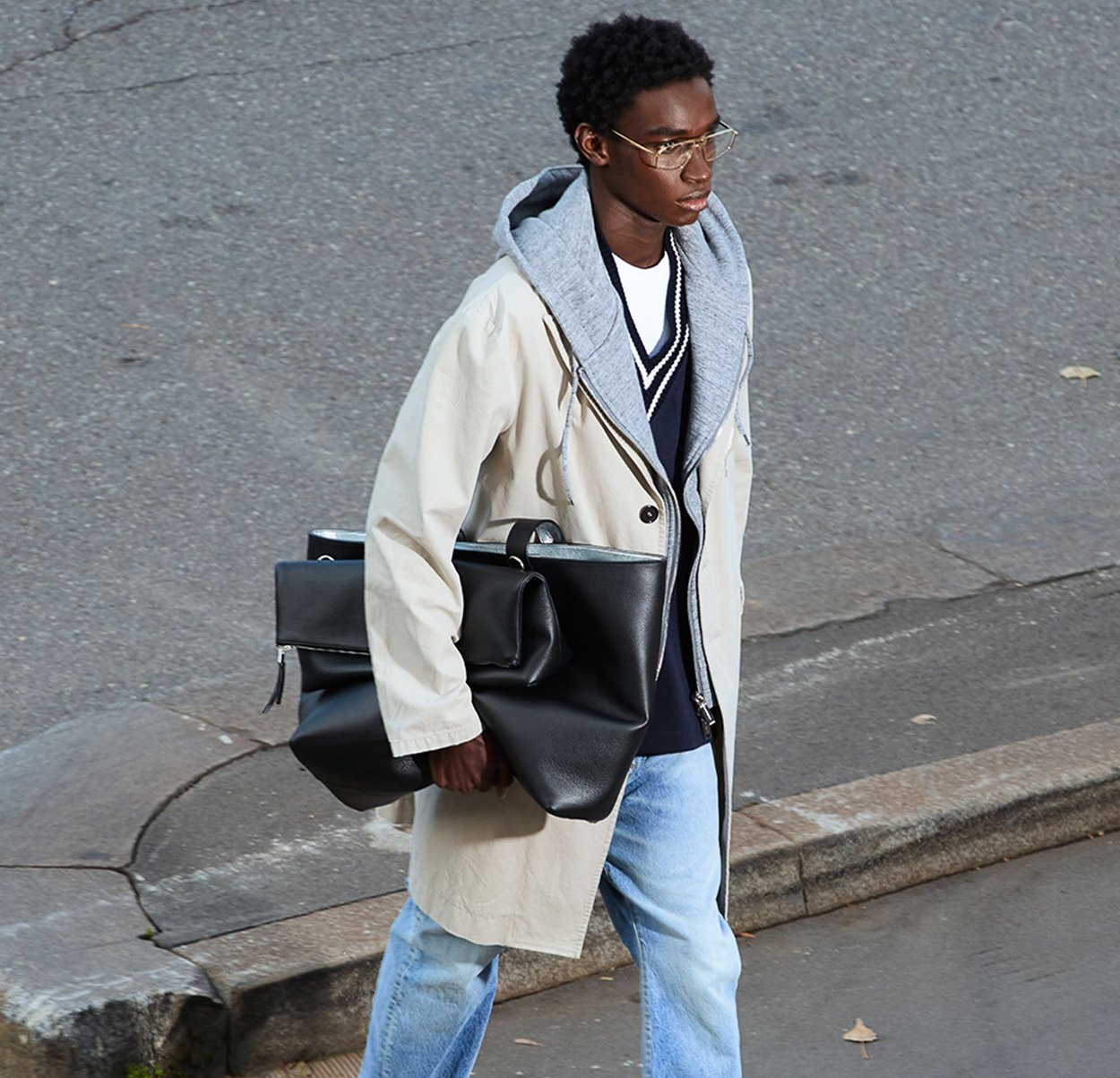 Beige Jacke mit grauem Kapuzenaufsatz, blauer Pullover mit weißen Akzenten, hellblaue Jeans und trägt eine große schwarze Tasche. Straßenszene.
