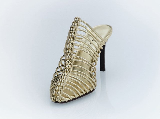 Ornate gold strappy high-heeled shoe showcasing a braided design at the front. It has an open back and a pointed toe. Enhanced by a smooth, metallic finish.
