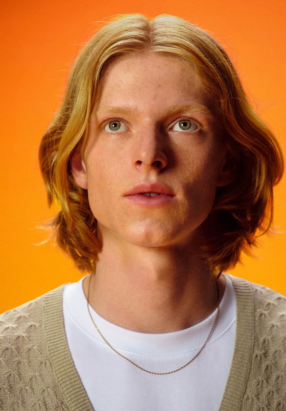 Person mit welligem, schulterlangem blonden Haar und heller Haut, trägt ein weisses T-Shirt und eine gestrickte beigen Strickjacke vor einem orangefarbenen Hintergrund.