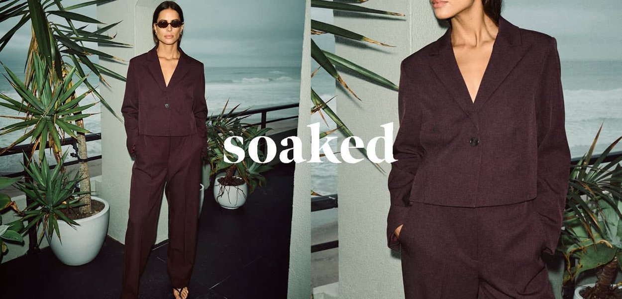 Dunkelviolette, taillierte, cropped Blazerjacke und dazu passende Hose mit glatter Textur, getragen mit schwarzen Sonnenbrillen, vor einem Küstenhintergrund.