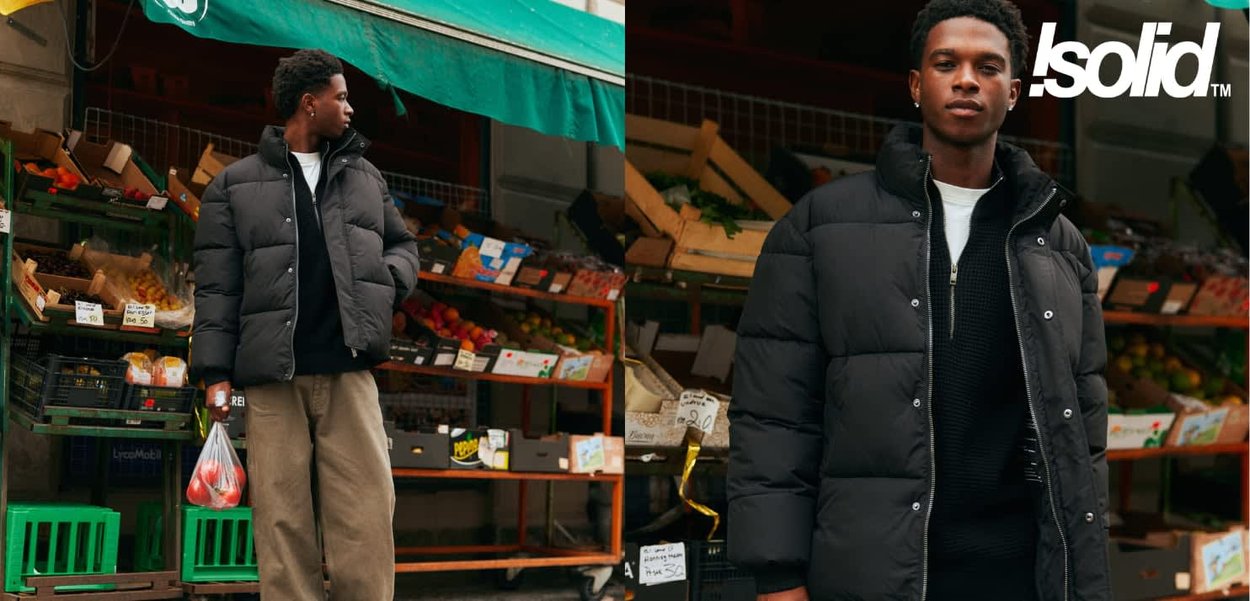 Giacca imbottita nera con design trapuntato, cerniera frontale e collo alto; indossata sopra un maglione scuro in un mercato della frutta con casse di legno.