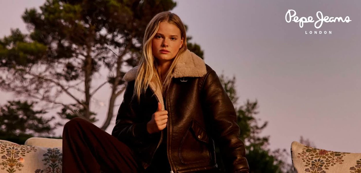 Veste en cuir marron avec un col en shearling beige, associée à un pantalon foncé. Le mannequin pose sur un canapé décoratif devant un décor naturel.