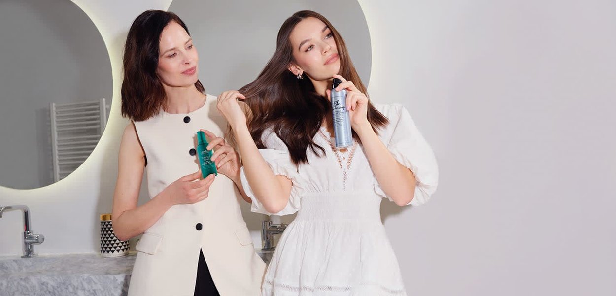 Dos mujeres en un baño aplicándose laca para el cabello, una con un vestido blanco y la otra con un chaleco beige sin mangas.