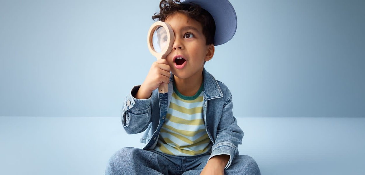 Mały chłopiec w pasiastym t-shircie i dżinsowej kurtce siedzi po turecku, zaskoczony, trzymając lupę przy jednym oku.