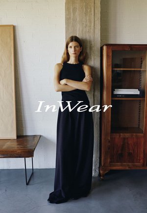 Frau in ärmellosem schwarzem Oberteil und langem schwarzem Rock steht mit verschränkten Armen in einem minimalistischen Raum mit Holzmöbeln und Betonsäule.