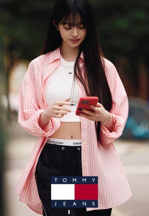 Jeune femme aux longs cheveux portant une chemise à rayures roses, un crop top blanc et un pantalon noir, utilisant un smartphone rouge en plein air.