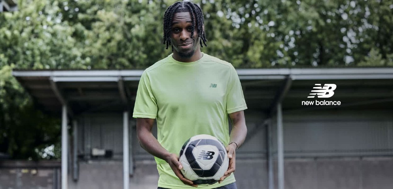 Groene New Balance sportshirt, korte mouwen, gestructureerde stof. Houdt een zwart-wit voetbal met logo vast, staande in de buitenlucht.
