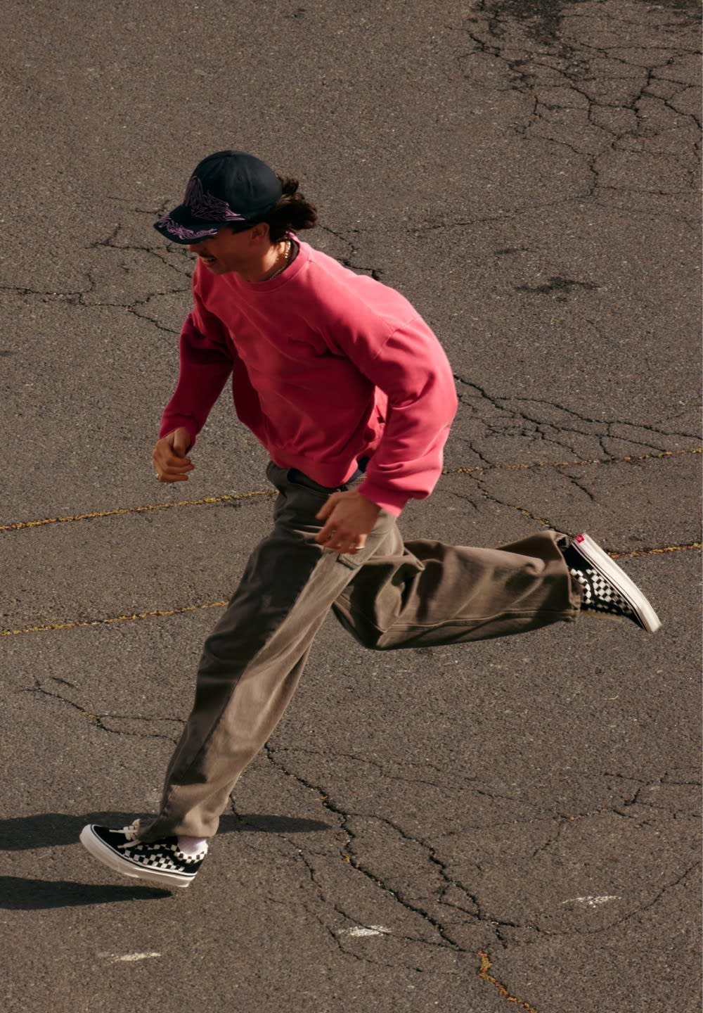 Tekmovalec v roza puloverju, sivih hlačah in črno-belo šahiranih supergakh, na razpokanem asfaltu. Pulover ima ohlapen krojni kroj in okrogel izrez.