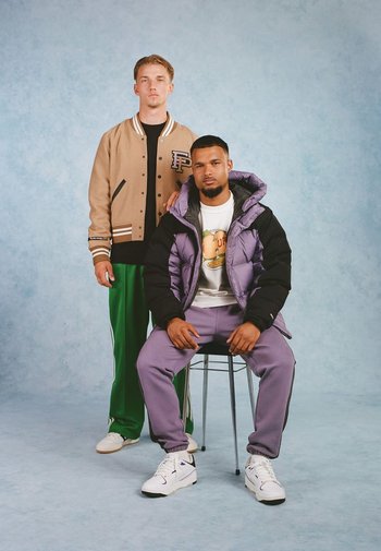 Twee mannen in casual outfits. De een draagt een bruine varsity jack, de ander een paarse pufferjack over een wit T-shirt en paarse broeken.