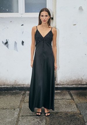 Vrouw staat buiten en draagt een lange zwarte satijnen jurk met dunne bandjes en kraaltjesdetails op het lijfje, zichtbare zwarte open-teenhakken.