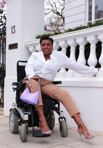 Elektrisk rullstol med svart ram och silverdetaljer, utrustad med kraftiga hjul. Person i vit skjorta, beige byxor och lavendelfärgad väska.