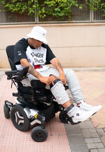 Elektrisk rullstol med en slimmad svart ram, stora gummihjul och justerbara armstöd. Användaren har på sig en vit bucket-hatt och avslappnade kläder.