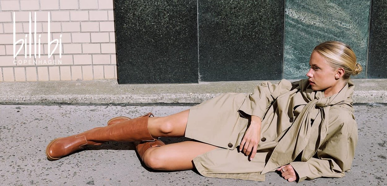 Trench coat beige avec un détail de ceinture, associé à des bottes en cuir marron jusqu'aux genoux. Fond de mur texturé dans des couleurs sourdes.