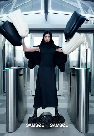 Kvinde i sort kjole og hætte står på elektrisk hoverboard og kaster sorte og hvide tasker inde gennem en moderne indgangsport af glas og metal.