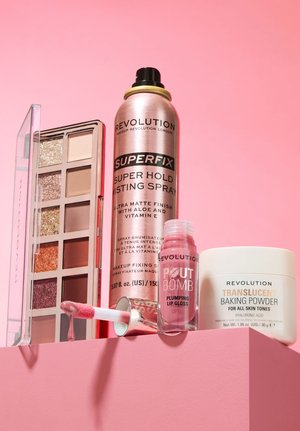 Palette di ombretti, spray fissativo, gloss labbra rosa con applicatore e cipria trasparente esposti su una superficie rosa con sfondo rosa.