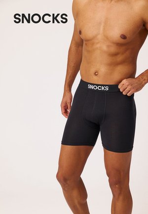 Skjorteløs mand iført sorte boxershorts med "SNOCKS" logo på linningen mod en ensfarvet baggrund.