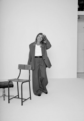 Grå oversized blazer og løse bukser over en hvid skjorte; står ved siden af en metalstol og en træpude på en lys baggrund.