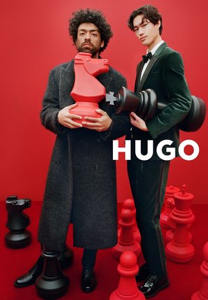 Due individui tengono pezzi da scacchi sovradimensionati rossi e neri contro uno sfondo rosso acceso. Un pezzo è un cavaliere stilizzato, l'altro un re.