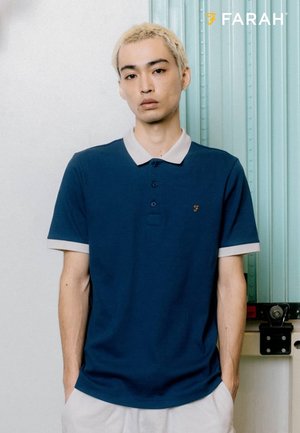 Giovane uomo con capelli biondi corti che indossa una polo blu con colletto e bordi delle maniche chiari, in piedi davanti a una parete e una porta chiara.