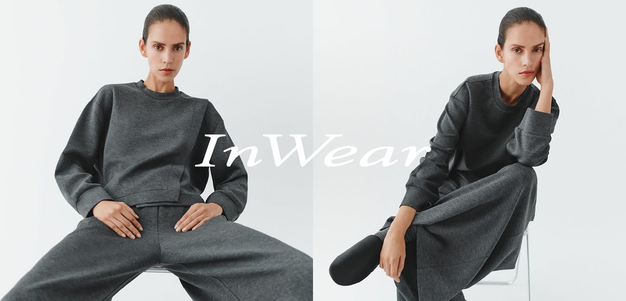 Grauer Pullover und weit geschnittene Hose aus weichem, strukturiertem Stoff, mit lockerem Sitz und schlichtem Ausschnitt. Logoschriftzug ist im Vordergrund zu sehen.