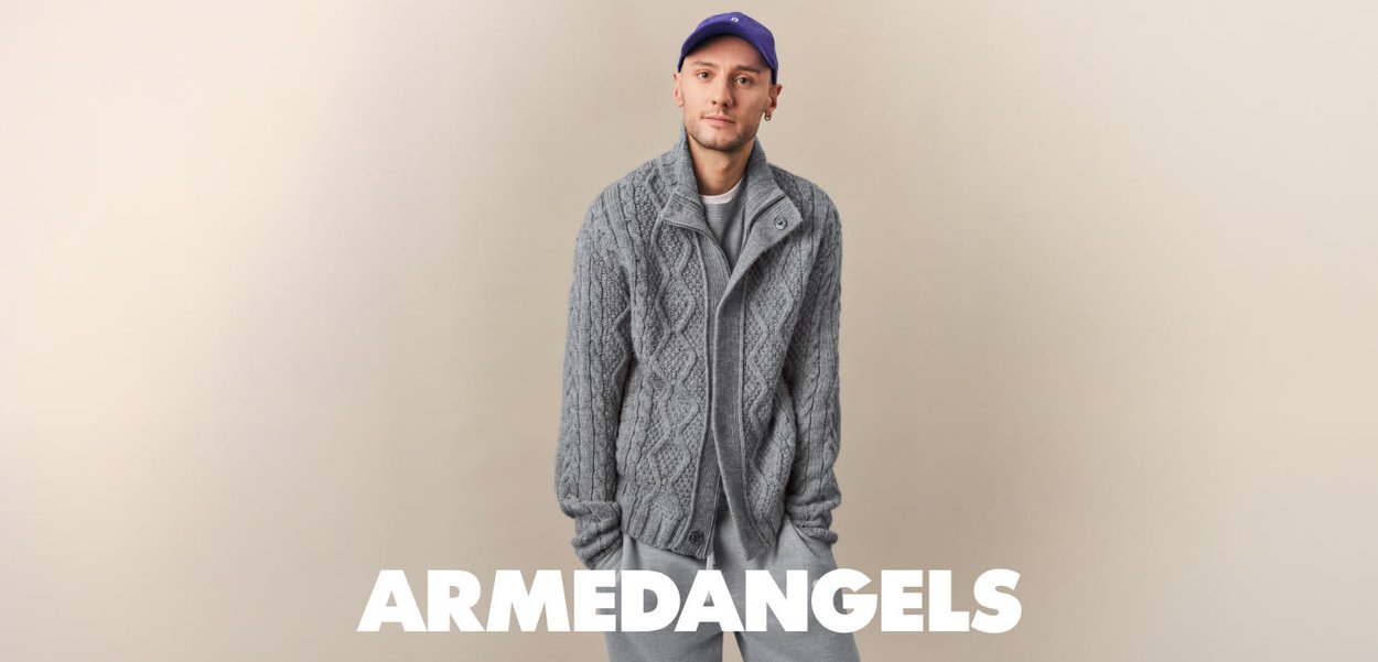 Gray knitted sweater with a cable design, featuring a tall collar and button closure, paired with gray sweatpants and a purple cap.