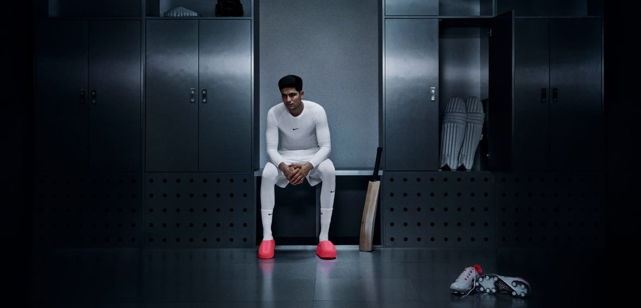 Athlet in enger weißer Kleidung mit Nike-Logo, sitzt auf einer Bank in einem schwach beleuchteten Umkleideraum. Helle pinke Schuhe an den Füßen, Cricketschläger in der Nähe.