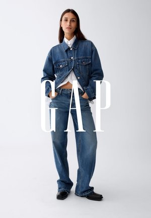 Donna in piedi con le mani nelle tasche, indossa una giacca di jeans corta, una camicia bianca, jeans a gamba larga e scarpe nere su sfondo bianco.