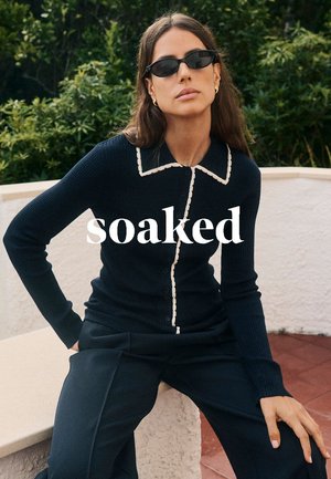 Marineblauer, gerippter Strickcardigan mit kontrastierendem weißen Kragen und Knöpfen, kombiniert mit dunkler Hose. Hintergrund zeigt grünes Laub.