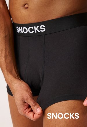 Nahaufnahme einer Person, die schwarze Snocks-Boxershorts trägt und mit einer Hand den Stoff am Oberschenkel anpasst.