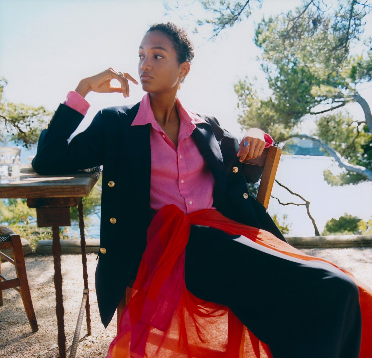 Mujer con blazer negro y camisa rosa sentada al aire libre junto a una mesa de madera, con árboles y agua visibles en el fondo.