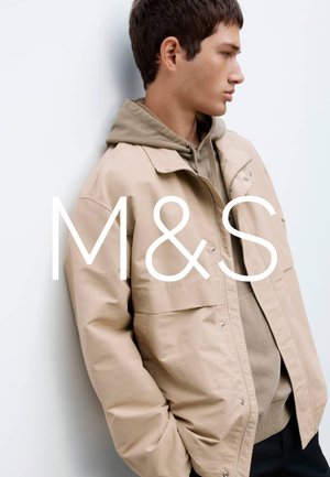 Beige, leichte Jacke mit Druckknöpfen und großer Fronttasche, getragen über einem khakifarbenen Hoodie. Modell posiert vor hellem Hintergrund.