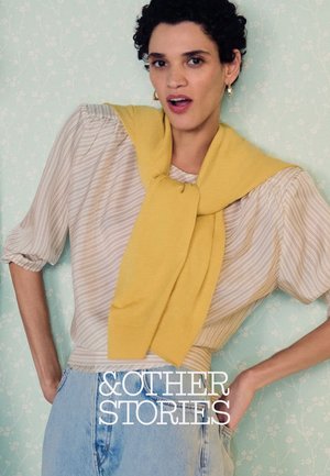 Frau mit kurzen lockigen Haaren trägt gestreifte Bluse, gelben Pullover über die Schultern gelegt und hoch taillierte hellblaue Jeans, posiert vor einer Blumenwand.