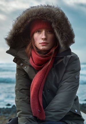 Young woman wearing a green parka with fur hood, red beanie, and red scarf, sitting outdoors near a body of water under a cloudy sky.