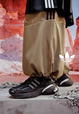 Person trägt beige weite Hose und schwarze Adidas-Sneaker und steht auf Beton vor rotem Felsenhintergrund.