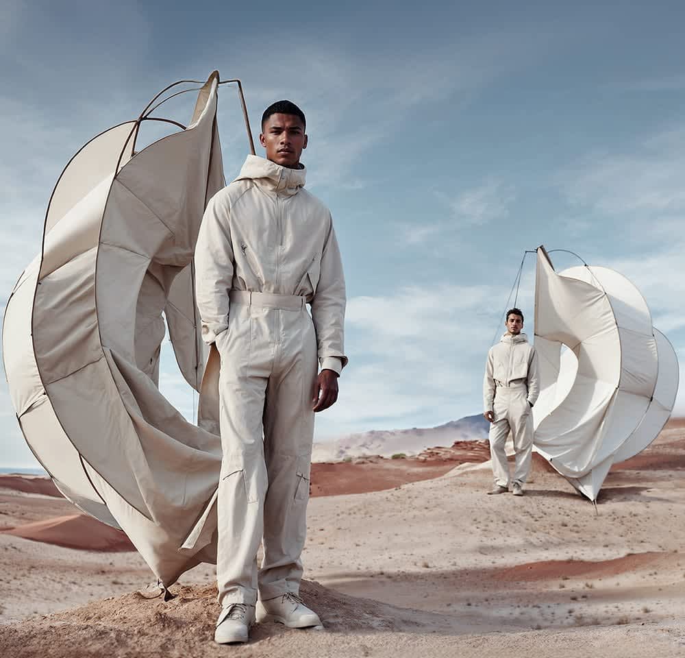 Beige outdoor jumpsuits with hoods, featuring large side pockets and matching footwear, near curved, white fabric structures on sandy terrain.