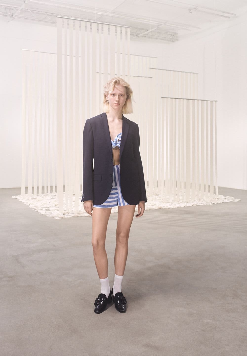 Black tailored blazer worn over a blue and white striped bikini top and matching shorts. Black shoes with buckles, white socks, neutral background.